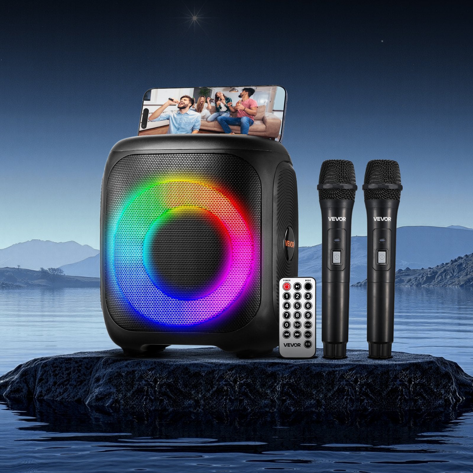 Portable speaker with RGB lighting, two microphones, and remote control on a rock by water with scenic background.