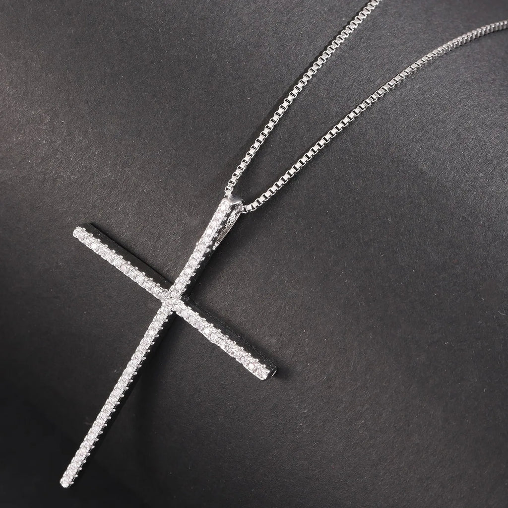 A silver cross pendant necklace paved with small clear stones, displayed on a textured black background.