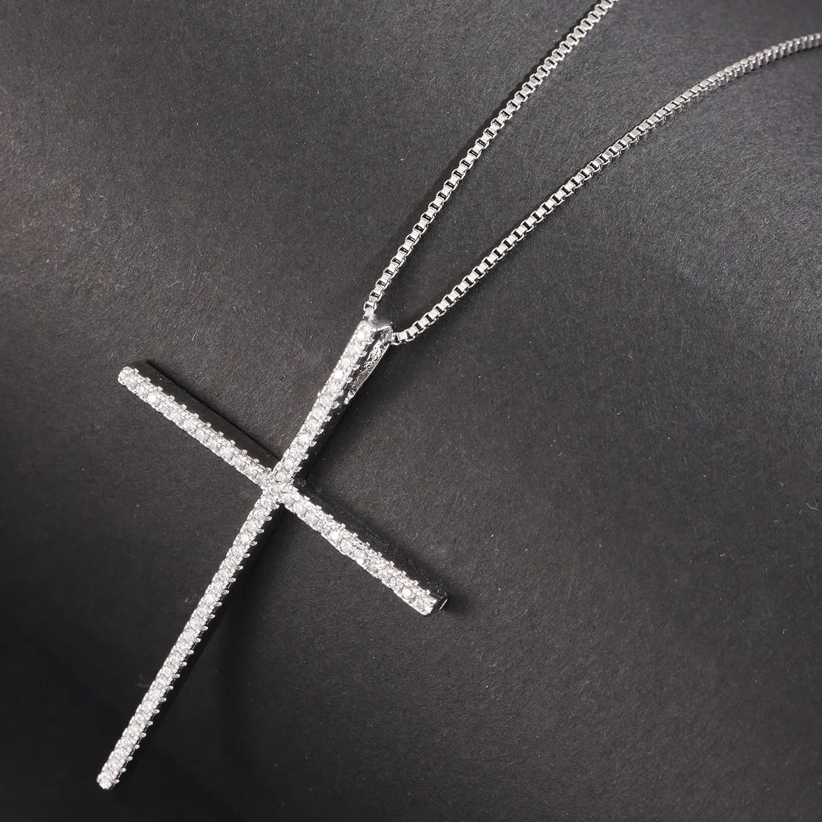 A silver cross pendant necklace paved with small clear stones, displayed on a textured black background.
