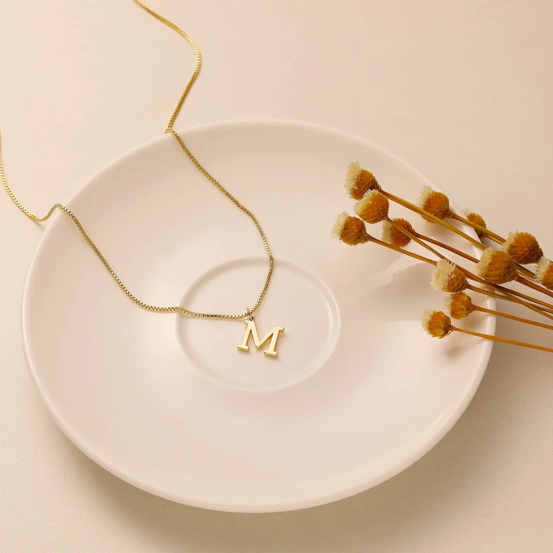 Gold necklace with 'M' pendant on a white plate with dried flowers
