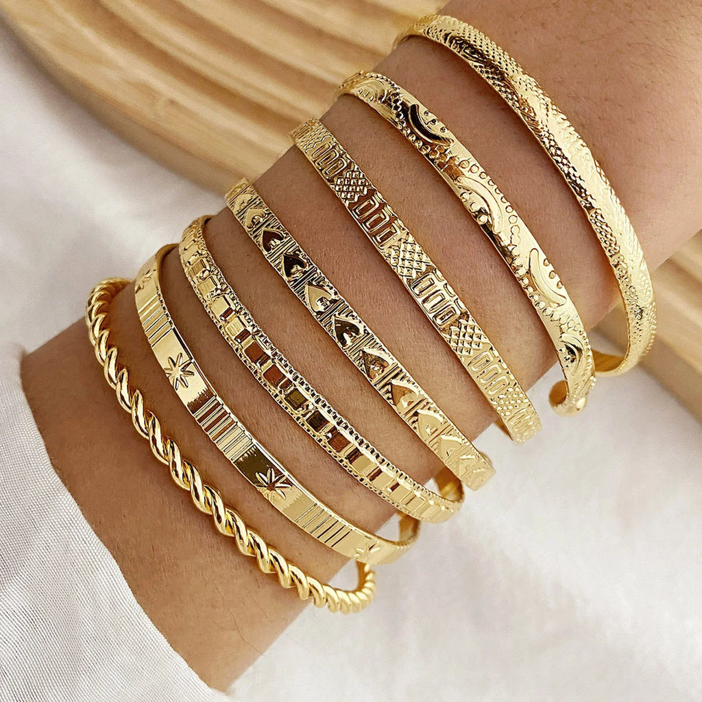 A close-up of a person's wrist wearing eight stacked gold bangles with different traditional Indian engravings and patterns, set against a soft white and beige fabric background.